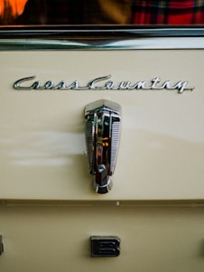 A close-up view of a vintage car with the logo 'Cross Country' prominently displayed above a metallic handle. The car is painted in a creamy beige color. There is a partial view of a plaid fabric and an orange object reflected in the background.
