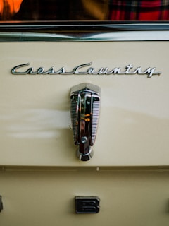 A close-up view of a vintage car with the logo 'Cross Country' prominently displayed above a metallic handle. The car is painted in a creamy beige color. There is a partial view of a plaid fabric and an orange object reflected in the background.