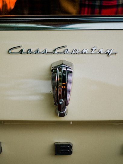 A close-up view of a vintage car with the logo 'Cross Country' prominently displayed above a metallic handle. The car is painted in a creamy beige color. There is a partial view of a plaid fabric and an orange object reflected in the background.