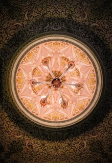 A beautifully ornate ceiling features intricate geometric patterns and floral designs in gold against a soft pink background. The centerpiece is a chandelier-like structure with multiple arms extending outwards, each adorned with decorative detailing. The ceiling is encircled by a richly patterned border filled with detailed motifs and floral elements.