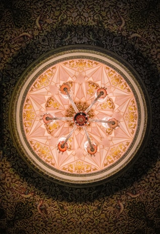 A beautifully ornate ceiling features intricate geometric patterns and floral designs in gold against a soft pink background. The centerpiece is a chandelier-like structure with multiple arms extending outwards, each adorned with decorative detailing. The ceiling is encircled by a richly patterned border filled with detailed motifs and floral elements.