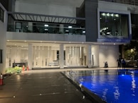 A modern building features a pool in the foreground with vibrant blue lighting. The exterior includes large glass windows, geometric wall patterns, and a deck area. Two people are standing near the pool, and there are exercise machines visible through the large windows on the upper floor.