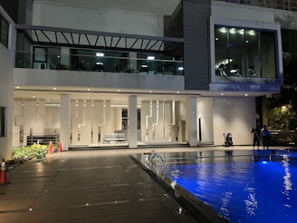 A modern building features a pool in the foreground with vibrant blue lighting. The exterior includes large glass windows, geometric wall patterns, and a deck area. Two people are standing near the pool, and there are exercise machines visible through the large windows on the upper floor.