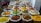 A vibrant display of a variety of spices in large white sacks, each with a scoop, arranged neatly in a market setting. The spices are of different colors and textures, including bright yellow, green, orange, brown, and reddish hues.