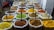A vibrant display of a variety of spices in large white sacks, each with a scoop, arranged neatly in a market setting. The spices are of different colors and textures, including bright yellow, green, orange, brown, and reddish hues.