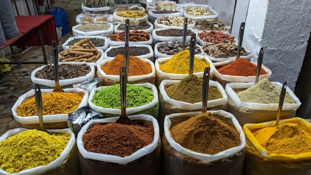 A vibrant display of a variety of spices in large white sacks, each with a scoop, arranged neatly in a market setting. The spices are of different colors and textures, including bright yellow, green, orange, brown, and reddish hues.
