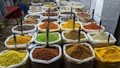 A vibrant display of a variety of spices in large white sacks, each with a scoop, arranged neatly in a market setting. The spices are of different colors and textures, including bright yellow, green, orange, brown, and reddish hues.