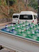 Large water bottles with blue and green caps are placed in the back of a white vehicle, possibly a truck. In the background, there are white vans parked on a road, with trees and a stone wall lining the side of the street.