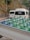 Large water bottles with blue and green caps are placed in the back of a white vehicle, possibly a truck. In the background, there are white vans parked on a road, with trees and a stone wall lining the side of the street.