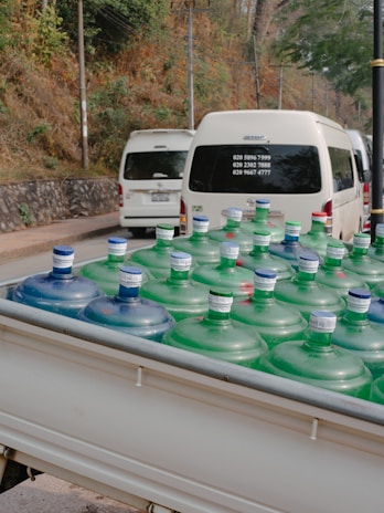 Large water bottles with blue and green caps are placed in the back of a white vehicle, possibly a truck. In the background, there are white vans parked on a road, with trees and a stone wall lining the side of the street.
