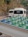 Large water bottles with blue and green caps are placed in the back of a white vehicle, possibly a truck. In the background, there are white vans parked on a road, with trees and a stone wall lining the side of the street.