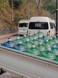 Large water bottles with blue and green caps are placed in the back of a white vehicle, possibly a truck. In the background, there are white vans parked on a road, with trees and a stone wall lining the side of the street.
