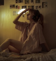 A person with headphones is sitting on a bed, wearing a striped shirt and holding one hand to their forehead. The room has dim lighting with a warm tone. Several small photos or pictures are displayed on the wall.