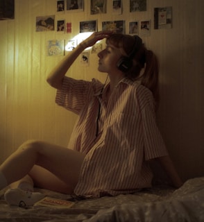 A person with headphones is sitting on a bed, wearing a striped shirt and holding one hand to their forehead. The room has dim lighting with a warm tone. Several small photos or pictures are displayed on the wall.