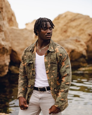 A person stands beside a body of water, wearing a camouflage jacket over a white shirt and light-colored pants. The rocky, earthy backdrop suggests a natural, rugged environment.