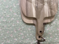 Two overlapping wooden cutting boards are placed on a fabric with a floral pattern. The boards are rectangular with rounded edges and have holes at the handle with leather strings attached. The fabric background is green with small white and pink flowers connected by delicate, vine-like stems.
