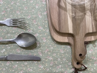 A set of silverware comprising a fork, spoon, and knife is placed on a green floral tablecloth. Adjacent to the silverware, two wooden cutting boards with leather strap handles are stacked together. A glass is visible on the top cutting board.