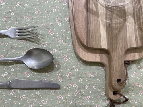 A set of silverware comprising a fork, spoon, and knife is placed on a green floral tablecloth. Adjacent to the silverware, two wooden cutting boards with leather strap handles are stacked together. A glass is visible on the top cutting board.