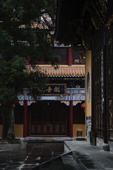 A traditional Asian architectural structure with ornate designs, featuring a roof with intricate patterns and red columns. The building is surrounded by lush greenery, and the atmosphere is serene and quiet, with soft lighting.