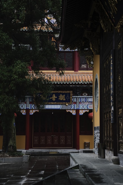 A traditional Asian architectural structure with ornate designs, featuring a roof with intricate patterns and red columns. The building is surrounded by lush greenery, and the atmosphere is serene and quiet, with soft lighting.