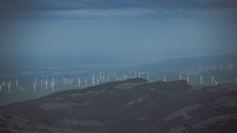 A vast landscape with rolling hills and numerous wind turbines scattered across the fields. The sky above is overcast with a hazy blue hue, and the terrain below consists of patches of green vegetation. The wind turbines are evenly spaced and line the horizon, indicating a large wind farm.
