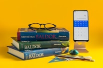 A stack of three mathematics textbooks is placed on a yellow surface, with a pair of black glasses resting on top. Nearby, a smartphone on a stand displays a calculator app showing the number pi. Various stationery items, including a pencil, eraser, and geometric tools, are also arranged nearby.