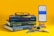 A stack of three mathematics textbooks is placed on a yellow surface, with a pair of black glasses resting on top. Nearby, a smartphone on a stand displays a calculator app showing the number pi. Various stationery items, including a pencil, eraser, and geometric tools, are also arranged nearby.