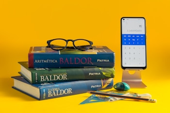 A stack of three mathematics textbooks is placed on a yellow surface, with a pair of black glasses resting on top. Nearby, a smartphone on a stand displays a calculator app showing the number pi. Various stationery items, including a pencil, eraser, and geometric tools, are also arranged nearby.