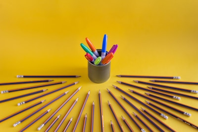 A collection of colorful markers is arranged in a container at the center with numerous pencils radiating outwards on a vibrant yellow background.
