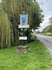 A signpost stands by a rural roadside, surrounded by lush greenery and overhanging trees, indicating the town of Louth with artistic designs. A brick base supports the wooden structure, accompanied by additional signage related to a local club. A red vehicle approaches in the distance on the paved road.
