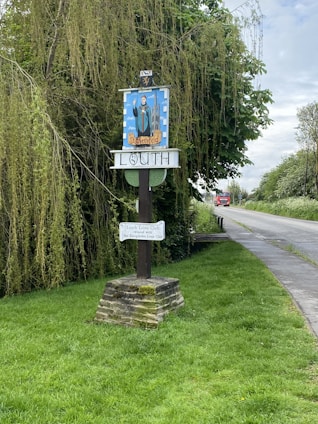 A signpost stands by a rural roadside, surrounded by lush greenery and overhanging trees, indicating the town of Louth with artistic designs. A brick base supports the wooden structure, accompanied by additional signage related to a local club. A red vehicle approaches in the distance on the paved road.