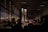 A dimly lit waiting area with rows of seats, occupied by several people sitting and facing forward. The area is illuminated by a warm, natural light, creating shadows and patterns on the walls and floor from slatted windows. The space feels contemplative, with an ambient, serene atmosphere.