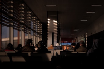 A dimly lit waiting area with rows of seats, occupied by several people sitting and facing forward. The area is illuminated by a warm, natural light, creating shadows and patterns on the walls and floor from slatted windows. The space feels contemplative, with an ambient, serene atmosphere.