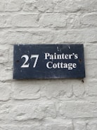 A dark blue rectangular sign with white lettering that reads '27 Painter's Cottage' is mounted on a light gray brick wall. The wall has a rough texture with visible bricks and mortar lines.