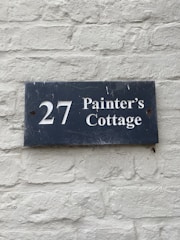 A dark blue rectangular sign with white lettering that reads '27 Painter's Cottage' is mounted on a light gray brick wall. The wall has a rough texture with visible bricks and mortar lines.