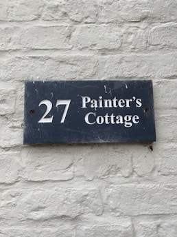 A dark blue rectangular sign with white lettering that reads '27 Painter's Cottage' is mounted on a light gray brick wall. The wall has a rough texture with visible bricks and mortar lines.
