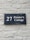 A dark blue rectangular sign with white lettering that reads '27 Painter's Cottage' is mounted on a light gray brick wall. The wall has a rough texture with visible bricks and mortar lines.