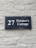 A dark blue rectangular sign with white lettering that reads '27 Painter's Cottage' is mounted on a light gray brick wall. The wall has a rough texture with visible bricks and mortar lines.