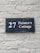 A dark blue rectangular sign with white lettering that reads '27 Painter's Cottage' is mounted on a light gray brick wall. The wall has a rough texture with visible bricks and mortar lines.