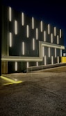 A modern architectural facade featuring a series of vertically aligned white fluorescent lights against a dark background. The lights create a striking pattern, and there is a series of geometric lines complementing the design, including a frame-like structure with ambient yellow lighting along the edges. The floor appears textured, likely concrete, with a subtle reflection of the lights. A touch of yellow can be seen towards the right, adding contrast to the overall design.