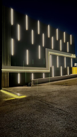 A modern architectural facade featuring a series of vertically aligned white fluorescent lights against a dark background. The lights create a striking pattern, and there is a series of geometric lines complementing the design, including a frame-like structure with ambient yellow lighting along the edges. The floor appears textured, likely concrete, with a subtle reflection of the lights. A touch of yellow can be seen towards the right, adding contrast to the overall design.