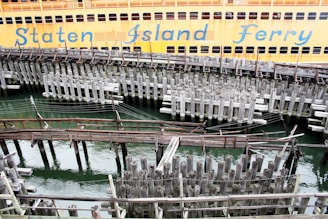 A large orange ferry is docked at the pier, with the words 'Staten Island Ferry' visible on its side. The foreground features a complex network of old, weathered wooden pilings and beams above the greenish water.