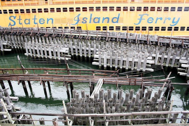 A large orange ferry is docked at the pier, with the words 'Staten Island Ferry' visible on its side. The foreground features a complex network of old, weathered wooden pilings and beams above the greenish water.