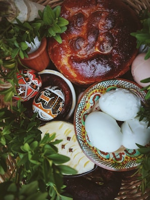 A festive arrangement featuring a braided loaf of bread with a shiny crust, colorful decorated eggs, white hard-boiled eggs in a patterned bowl, and a creamy cheese with raisins or cloves. The scene is surrounded by vibrant green leaves and rustic elements, suggesting a celebratory or cultural theme.