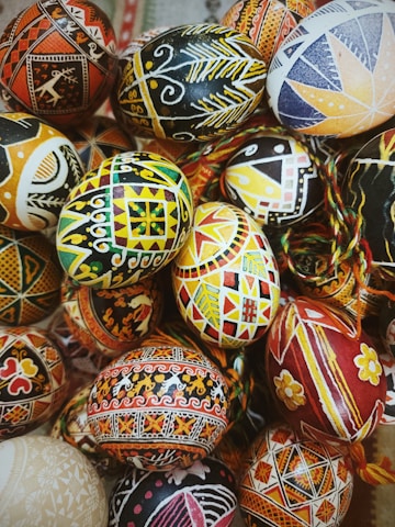 A collection of intricately decorated eggs, featuring a variety of vibrant, geometric, and floral patterns. The designs are highly detailed, with a mix of colors including black, yellow, red, and orange, creating a striking and artistic visual. The eggs are arranged closely together, emphasizing their diversity in patterns and design.