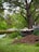 A garden setting with a large tree in the background. In the foreground, a pile of soil is next to a wheelbarrow filled with sand or soil. There is greenery all around, including grass and plants, with a white picket fence visible in the distance.
