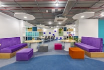 A modern office space featuring colorful seating and tables. The area includes purple couches and padded stools in orange and pink, set against a background of open workstations. Overhead lighting is minimal and geometric, while greenery adds a touch of nature. The decor is vibrant and inviting, with a mix of textures and colors.