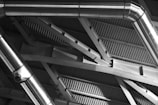 Metal ducts and beams form an intricate pattern against the backdrop of a corrugated metal ceiling. The shiny surfaces of the ducts reflect light, creating a play of highlights and shadows across the architectural structure.