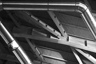 Metal ducts and beams form an intricate pattern against the backdrop of a corrugated metal ceiling. The shiny surfaces of the ducts reflect light, creating a play of highlights and shadows across the architectural structure.