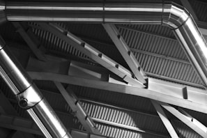 Metal ducts and beams form an intricate pattern against the backdrop of a corrugated metal ceiling. The shiny surfaces of the ducts reflect light, creating a play of highlights and shadows across the architectural structure.
